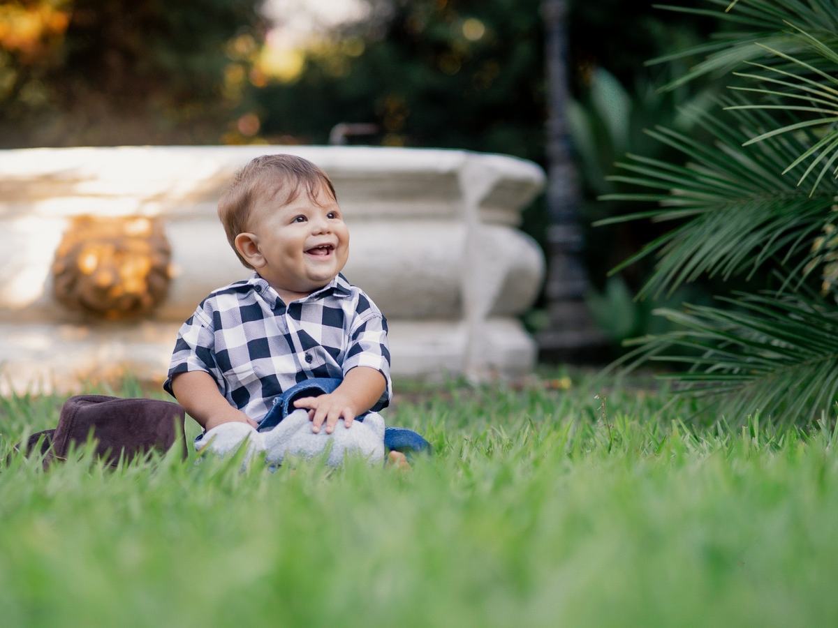Criança sentada na grama do sítio, sorrindo, com o jardim e o chafariz ao fundo no fim da tarde.