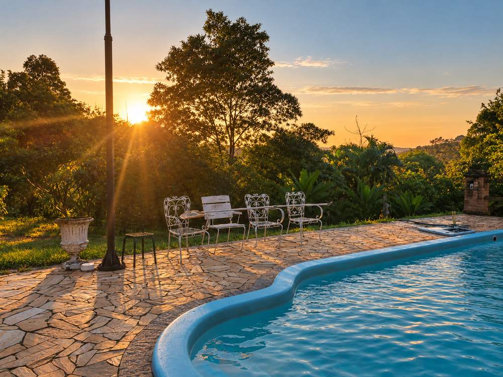 O fim de tarde na beira da piscina, com o sol baixando atrás das árvores e as cadeiras de ferro no deck de pedra.